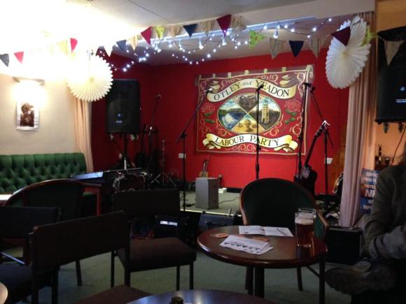 A few tables and chairs in front of a cosy-looking small stage with fairly lights and decorations. On the stage are 5 microphones and PA speakers, guitars and a violin, to the side is a mixing desk. On the red back wall of the stage is a painted mural of Otley and Yeadon Labour Party reminiscent of an old marching banner, red and gold, with roses and four images: the Otley clock tower, a field of cut corn, a weir in a river, a printing press.